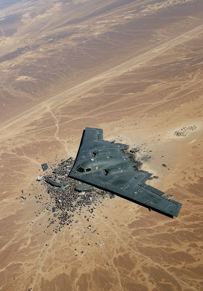 The image depicts a military aircraft, specifically a B-2 Spirit Stealth Bomber, lying in a desert environment. The aircraft appears to be damaged or destroyed, as debris is scattered around the crash site. The surrounding landscape is barren with sand dunes, indicating a remote and arid region. The image likely conveys a narrative of military conflict or the aftermath of a mission gone wrong.