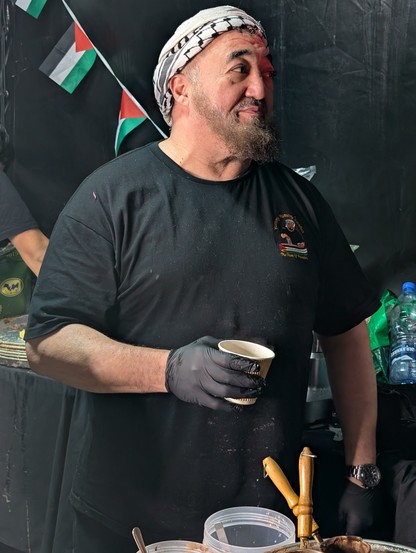 A solid Palestinian dude with a wry smile, holding a cup of Arabic coffee, wearing a kiffeyah and a t-shirt with his name and logo stitched in, and looking like a total baller