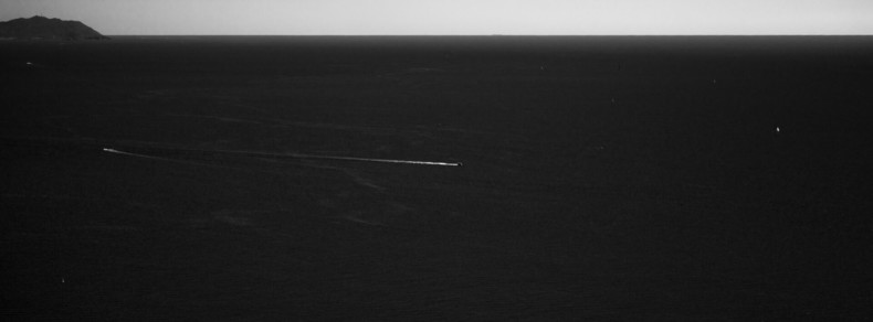 Aerial view of a vast, dark ocean with a small island visible on the far left horizon. A thin, bright line cuts across the water, likely indicating a distant boat's wake. The sky above is a lighter shade, contrasting with the dark sea below. Minimal visual elements contribute to the expansive and open landscape.