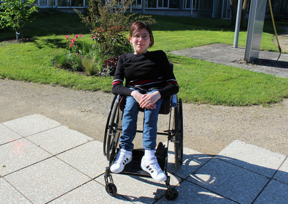 Eine junge Frau sitzt in einem Rollstuhl vor Wiese und Blumenbeet und lächelt in die Kamera.