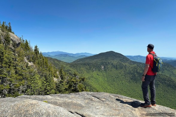 A hiker stands on a rugged rock ledge, gazing out over an expanse of forested mountains. They wear a red shirt, dark pants, a cap, and a backpack. The landscape stretches into the distance, with lush greenery covering the slopes under a clear blue sky. The scene conveys a strong sense of adventure, solitude, and connection to nature.