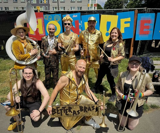 A group of musicians poses outdoors, all wearing shiny gold outfits. They hold various brass instruments, including trumpets and a trombone. A sign in the foreground reads "BETTER AS BRASS" with colorful graphics. The setting appears festive and vibrant.