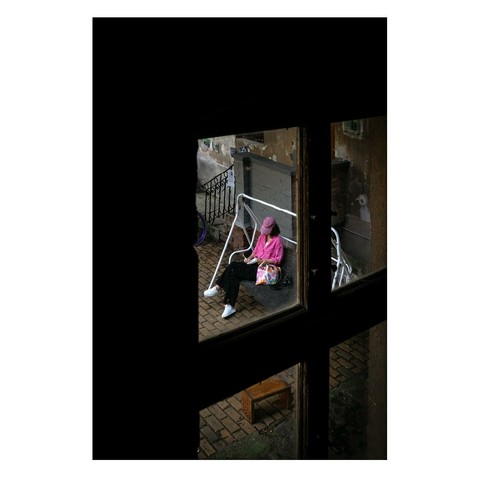 The picture was taken from the window of the stairwell of an old apartment building in the city center. In the courtyard, a woman wearing a pink top was sitting on a white swing bed and writing in a notebook. On the left side of the picture, you can see the darkness of the stairwell and the frame of the window through which the photo was taken.