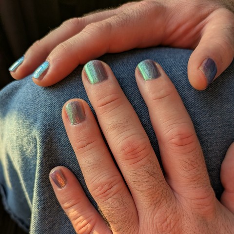 Close up on two hands sitting on a denim-covered knee, showing off fingernails. The colors of the nails vary in a range between teal, some purples, and a coppery gleam.