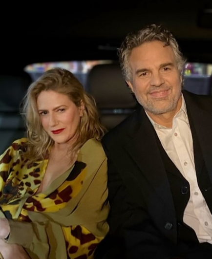 Mark Ruffalo y su mujer sentados en un vehículo, ambos sonriendo mientras visten ropa elegante. El entorno es oscuro, pero se perciben luces difusas al fondo.