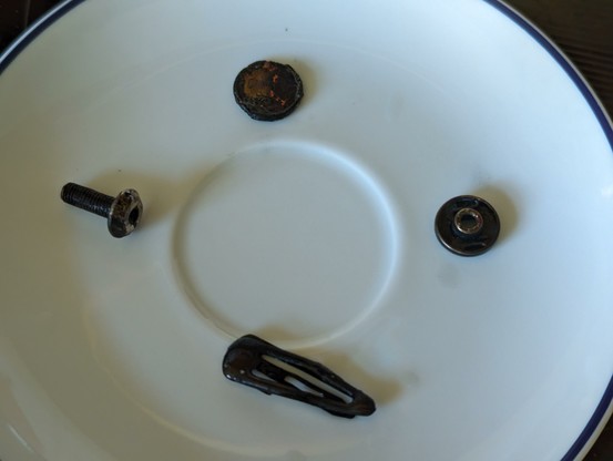 Auf einem Tellerchel liegen vier Schwarze Teile. Eine Schraube, ein Knopf, eine Haarspange und ein (vmtl.) Centstück