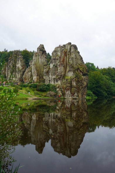 A wide, eye-level shot on an overcast day captures the Externsteine rock formation, a natural monument in North Rhine-Westphalia, Germany. Three imposing, weathered sandstone pillars, richly textured with fissures and patches of moss, rise majestically from a lush green landscape. The largest rock, on the right, features carved openings resembling windows or caves. To the left, a smaller pillar stands slightly behind the main formation, with a third, even smaller one partially visible further left, nestled amongst trees.
The rocks are reflected with striking clarity in the still, dark water of a lake in the foreground, creating a symmetrical visual effect. The banks of the lake are verdant, with green grass and foliage. A small pathway or boardwalk with a wooden fence is visible at the base of the rocks, suggesting accessibility for visitors. The overall scene conveys a sense of natural grandeur and historical significance.
