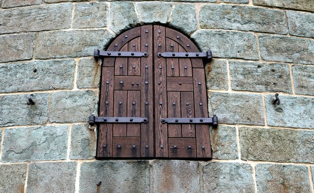 Close up photograph of dark brown wooden shutters, closed on a window in a grey stone wall. The shutters have four black metal hinges and rows of nails.