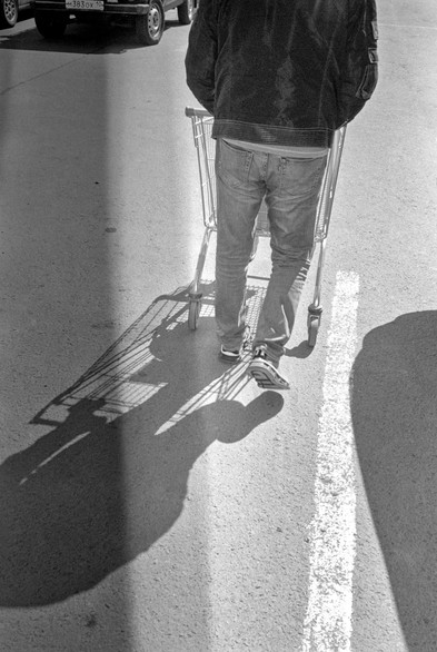 On a monochrome photo a man is walking with a grocery cart and casts a contrasting shadow.