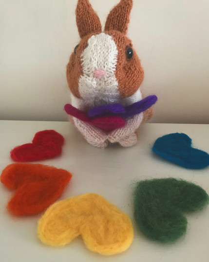 A knitted white and brown bunny holding a pink and purple needle-felted heart and surrounded by more needle felted hearts in red, orange, yellow, green, and blue