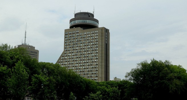 Photograph of a modern building built in grey concrete, more than twenty storey high, behind tall green trees. The building has rows of very regular rectangular windows and a dark brown circular structure on top with windows all around. It is in fact a rotating restaurant with a 360 degree view of the surroundings.