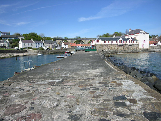 Lower Largo. The image shows a view along a broad stone pier to a village at its landward end. There is a large beige building on the right and houses on the left, with three arches of a railway viaduct visible in the centre of the frame. The sky is largely blue.