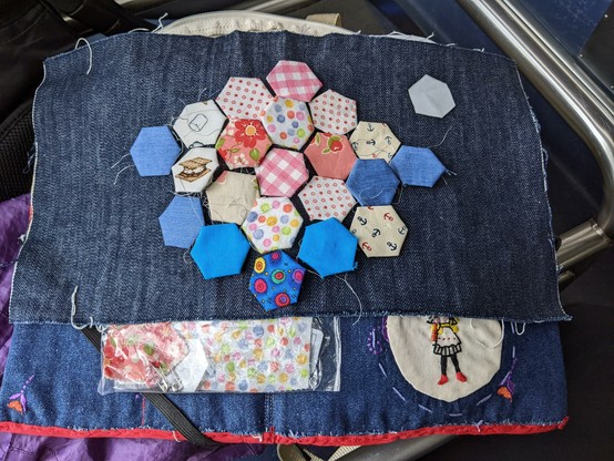 Laid out on an oblong piece of dark blue denim are twenty-one hexagonal fabric patches of various patterns and hues, basted with dull silver thread to paper templates. One template rests to the side of the patches.