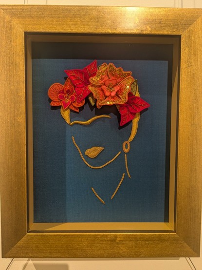 Needlework art piece showing a simplified image of Friday Kahlo.  Just a line for the face shape and a solid line for her monobrow.  Then rally vibrant red and orange flowers in her hair.