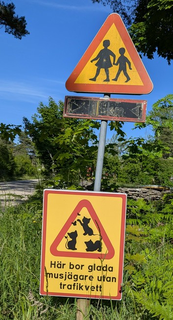 A photo of a rural road in Gotland, Sweden, with a segment of grey road to the left, green trees, grasses, and ferns under a blue sky. In the middle is a sign post with 2 signs. The top is a yellow triangle with a red border, and in the middle are the black silhouettes of a girl and a smaller boy walking hand in hand.

Beneath that is a square yellow sign with a red outline, and in the middle, surrounded by a red triangle , are the silhouettes of 3 kittens playing with butterflies.  Below are three lines of text in Swedish, which translates to "Here live happy mouse hunters lacking traffic knowledge".