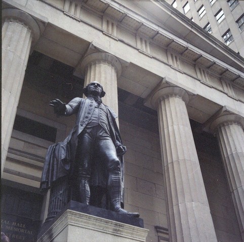 The statue of George Washington on Wall Street, New York. It's a dark grey bronze statue of a standing man in 18th century clothes in front of a white classical building with columns and a plinth, shot from a lowish angle.