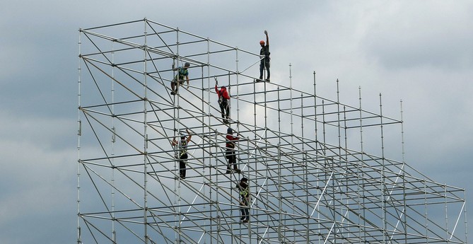 Photograph of six scaffolders working high up in a scaffolding metal structure, all wearing security helmets and harnesses, against a blue grey cloudy sky. Five of the workers are working inside the structure. The sixth worker is standing up on an horizontal bar at the very top, looking and waving at the photographer with his left arm in the air.