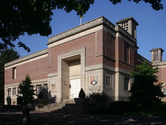 The image appears to show a building characterized by a combination of brick and stone architecture. It features a prominent entrance with steps leading up to double doors, and there are decorative elements and possibly coat of arms or emblems on the façade. The setting is outdoors with trees and clear blue skies visible, suggesting a well-maintained landscape. The style may indicate it is an institutional or historical building.

Image Credits: Wikimedia / GavinWarrins / Public domain