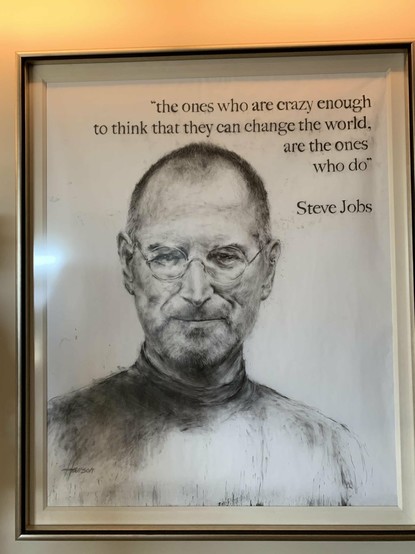 Framed portrait drawing of an older man with round glasses, clad in a simple high-collar shirt and showing a restrained smile. An attributive quote above reads: “the ones who are crazy enough to think that they can change the world, are the ones who do” - Steve Jobs. The drawing is signed by the artist at the bottom. A subtle warm light casts on the top edge of the frame.