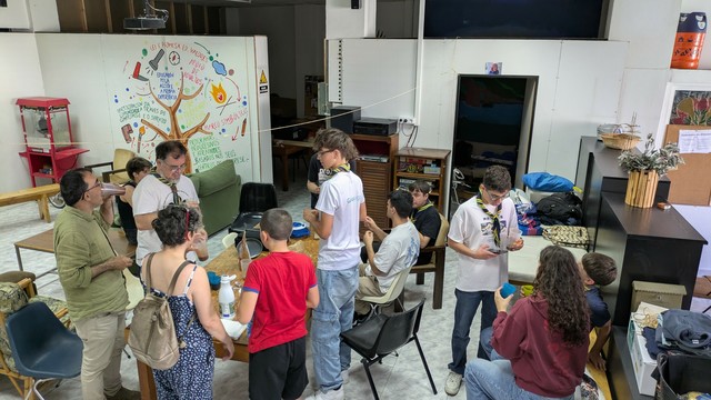 Un grupo numeroso de jóvenes, se encuentran en una sala comunitaria. Hay mesas con objetos sobre ellas, y en la pared hay un mural con dibujos coloridos relacionados con la naturaleza y actividades al aire libre. Están tomando chocolate.