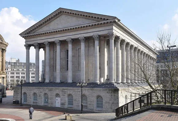 The image depicts a large neoclassical building characterized by its numerous tall columns and a prominent, grand façade. The architectural style is typically associated with ancient Greek and Roman designs. The building appears to be situated in a public square, surrounded by other structures, and features a clean, well-maintained exterior. The sky is partly cloudy, contributing to a bright atmosphere.

Image Credits: Wikimedia / Very Quiet / CC BY-SA 2.0
