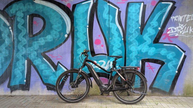Ein schwarzes Haibike Trekking steht vor einem Graffiti, das die Buchstaben DRUK zeigt. Was auch immer das bedeuten mag ;-) 
Die Schrift ist Türkis, der HIntergrund in lila.