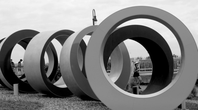 Black and white photograph of an art installation named "Des perles en mémoire" (Remembering Through Beads, 2024) by Huron-Wendat artist named Ludovic Boney, in reference to wampum belts made out of beads by members of the First Nation he is a member of, for political and trading alliances. The sculpture is made of six giant steel rings, not quite aligned and turning slightly. On the left side through one of the ring, two people can be seen walking. On the right side, a woman running can be seen through two of the rings.