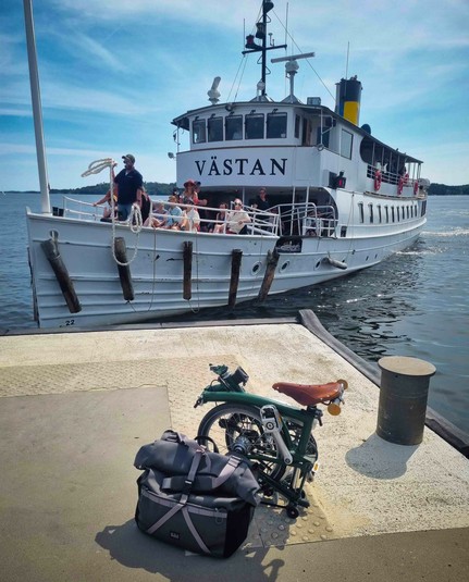 A folded folding bike with a bag on a landing bridge in the foreground. A small ferry boat just landing in the background. Blue sky.