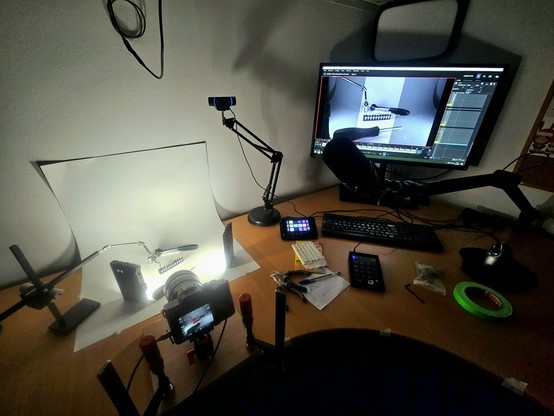 Foto eines rotbraunen Schreibtischs, auf dem allerlei Computertechnik zum Livestreamem sowie einige Kamera, Klebepads, Werkzeug, zwei kleine Lichter und ein verstellbarer Metallarm ("Rig") steht. Alles ist um eine Reihe Werkzeug-Bits drapiert, die von der Kamera auch auf einen hinten erkennbaren Bildschirm übertragen werden.