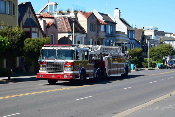 A red and white fire truck, labeled "San Francisco Fire Dept. 15," is driving down a street lined with multi-story houses featuring various architectural styles. The street appears mostly deserted and the sky is clear blue.
