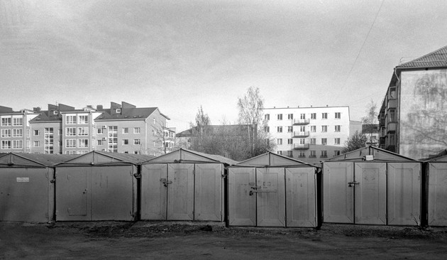 The monochrome photograph shows a row of metal old garages. High-rise buildings are visible in the far background.