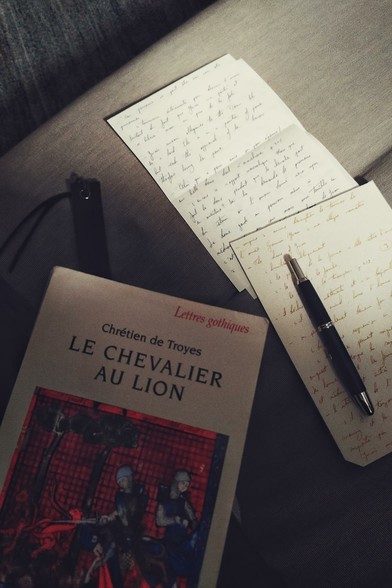 A vertical colour photograph of a book 'Le Chevalier au Lion' by Chrétien de Troyes rests on a grey sofa. Next to it are a few loose leaves of paper with handwritten notes. On the paper is a fountain pen.