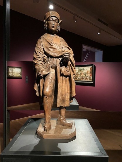 Statue en bois clair d’un homme portant une longue tunique, dont il relève un pan pour montrer une grande plaie que sa cuisse droite ainsi exposée.