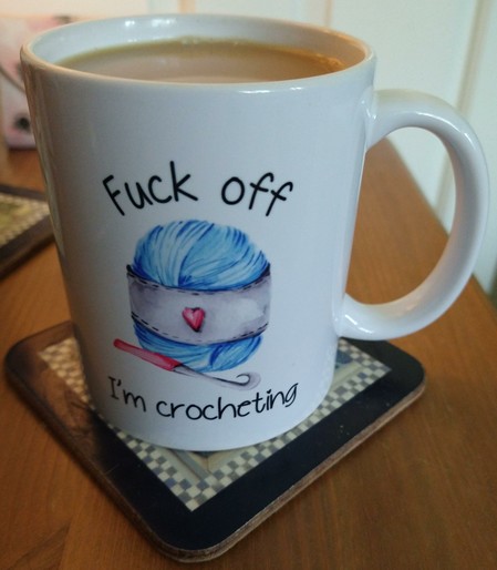Mug of tea on a bedside cabinet with the words 'fuck off I'm crocheting' surrounding a ball of blue yarn and a crochet hook