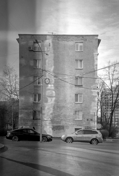 Monochrome photo shows a five-story house with a lot of glare reflected from the windows of a neighboring building. Two cars are parked in front of the house.