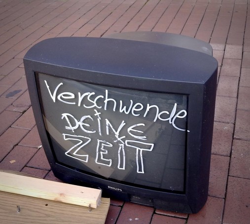 Zu sehen ist ein ausrangierter Röhrenfernseher der Marke Philips, der auf einem roten Pflastersteinboden steht. Auf dem Bildschirm steht in weißer, handgeschriebener Schrift der provokante Satz: „Verschwende deine Zeit“. Das Gerät ist offensichtlich defekt oder entsorgt worden. Neben dem Fernseher liegt ein Stück Holz mit Schrauben – möglicherweise Teil eines alten Möbelstücks. Die Inszenierung wirkt wie eine stille Anklage gegen den passiven Medienkonsum und verweist symbolisch auf die gesellschaftliche Zeitverschwendung durch inhaltsleeres Fernsehen und Boulevardformate.

Image Description (English):
The image shows a discarded Philips CRT television placed on a red brick pavement. Written in bold white handwriting across the screen is the provocative phrase: “Waste your time.” The TV appears broken or thrown away. Next to it lies a wooden board with screws, possibly from an old piece of furniture. The scene presents a symbolic critique of passive media consumption and alludes to the societal waste of time through empty television content and tabloid formats.🖖
