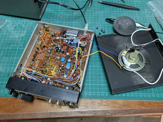 A CB radio with the cover taken off. The insides show a PCB, lots of wires and a speaker on a desk