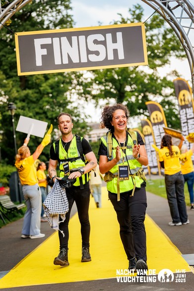 Twee deelnemers, zichtbaar vermoeid maar blij, lopen onder een grote boog met het woord "FINISH" tijdens de Nacht van de Vluchteling 2025 op 14/15 juni. Ze dragen gele veiligheidshesjes met startnummers, en worden toegejuicht door vrijwilligers in gele shirts. Op de achtergrond zijn vlaggen en borden met het logo van het evenement zichtbaar.