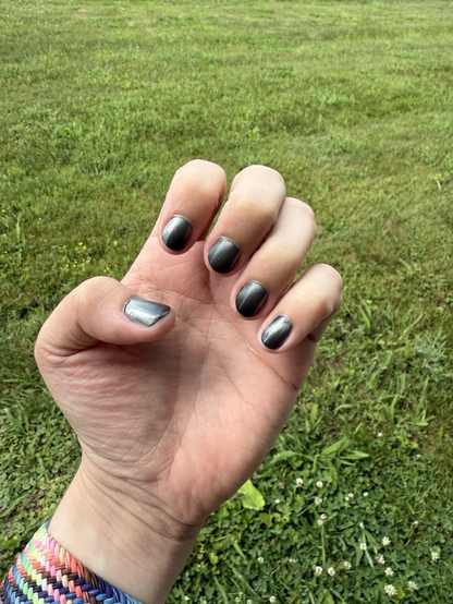 Photo of a white man’s hand shaped in a claw to show off metallic grey and black nail polish in the morning sunlight. Wrist has a woven, rainbow Pride colored watch band. Background is a freshly cut green lawn.