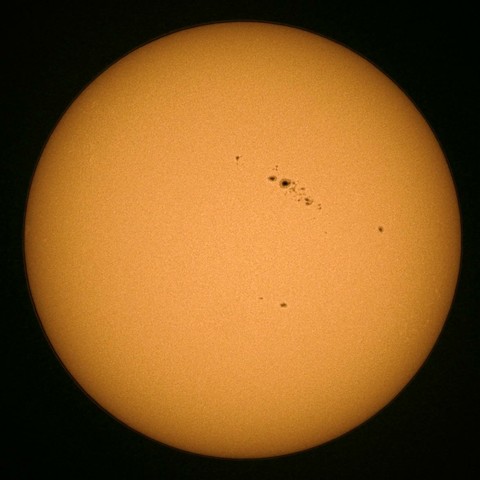 A close-up image of the Sun, predominantly orange with several sunspots visible on its surface.