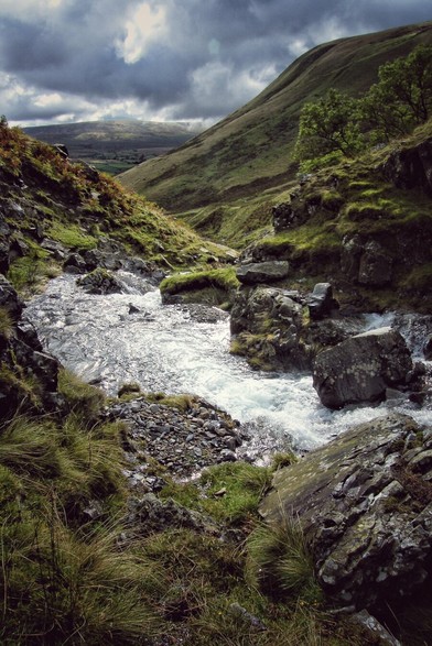 A dramatic view of a rocky stream cascading through a green valley near Cautley Spout in the Lake District. The water is bright and foamy, surrounded by dark, mossy rocks and lush green grass on the banks. Steep, grassy hills rise on either side under a moody, overcast sky with patches of bright light breaking through the clouds.