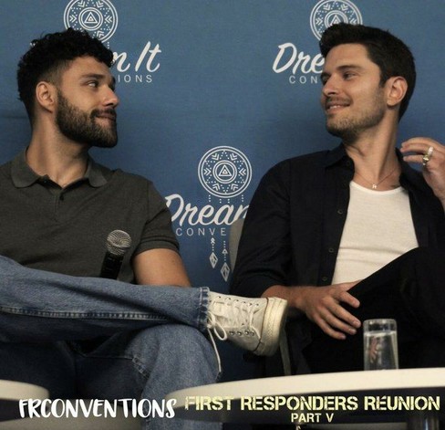 Rafael Silva (Carlos) and Ronan Rubinstein (T.K.) sitting next to each other on stage of the First Responders Reunion Convention V in France. They look at each other and it seems like they're communicating only with their eyes.