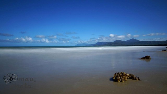 Sandy beach with a calm sea under a clear blue sky. Mountains are visible on the horizon, and small patches of seaweed are scattered across the wet sand in the foreground. Sparse clouds dot the sky.