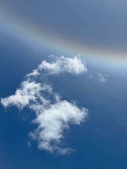 A clear blue sky with wispy white clouds scattered throughout. A subtle upside down rainbow gradient in the upper sky section of such a halo around the Sun.