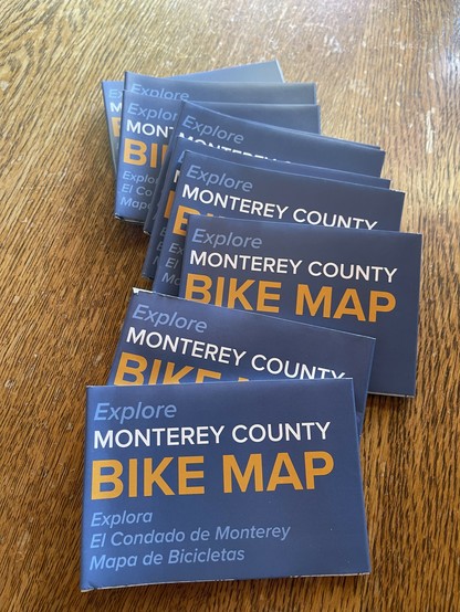 Some folded bike maps are on a wood tabletop. 

Text in English and Spanish, states: “Explore Monterey County - Bike Map.”