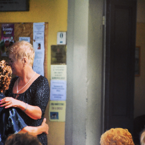 Two women dance together while others are watching. A yellow wall with many paper signs on it is behind them.