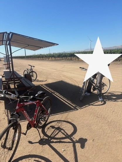 A man sits on a Giant hybrid bicycle in the Salinas Valley. Near him are two other bicycles, parked next to a shaded picnic table used by farm workers.