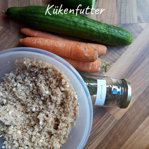 Auf einer Arbeitsplatte liegen drei dicke Möhren, eine Gurke und ein kleines Glas, nur noch spärlich gefüllt mit grau-grünen Samen und "Brennnessel" beschriftet, neben einer Schüssel, in der eingeweichter Couscous mit Haferflocken vermischt ist.