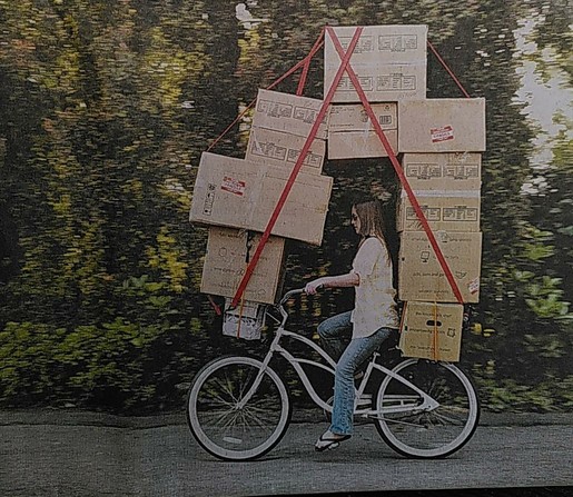Das Foto zeigt eine Radfahrerin, die mit neun Umzugskartons unterwegs ist. Die Szene demonstriert die billigstmögliche Art des Umzugs.
Die Anordnung der Kartons im Bogen vom Gepäckträger über die Fahrerin bis zum Fahrradkorb vorne sieht dank den Spanngurten stabil aus, ist aber nur als Spaß gemeint. Denn einerseits darf das Gewicht der Kartons in dieser Konstruktion nicht recht groß sein, andererseits wird die Sicht der Person auf dem Rad deutlich eingeschränkt.

Quelle: Titelbild der taz vom 19.6.2025 zum verlinkten Kommentar "Steigende Wohnkosten"