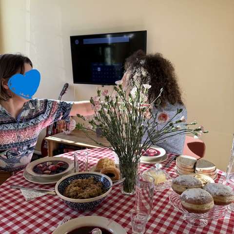 A table covered in red and white cloth, bowls of pierogi (polish dumplings) in red soup. A bowl filled with vegetables, and a plate with three donuts. A colourless vase in the middle, filled with dainty slightly pink flowers. Photo taken from one side, and on the other side two people are sitting down by the table. In the background is a pale yellow wall and a TV on the wall.